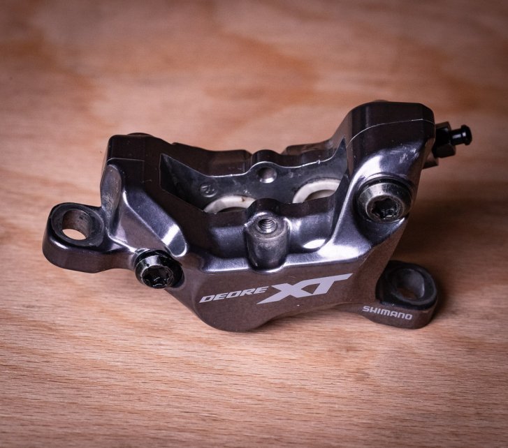 Una pinza de freno Shimano XT de 4 pistones descansa sobre una superficie de madera de haya. En su interior se distinguen claramente los pistones cerámicos de color claro.