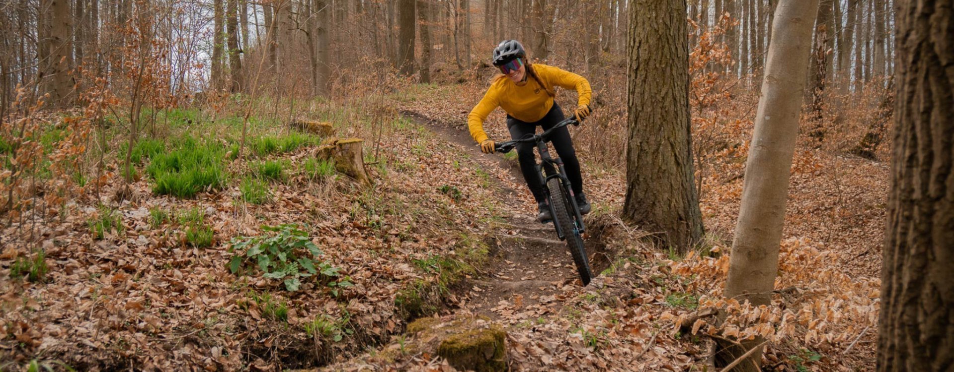 Mujer con maillot amarillo de MTB, casco negro y gafas de colores sonríe y señala su maillot mientras está sentada en una bicicleta de montaña en un bosque otoñal.