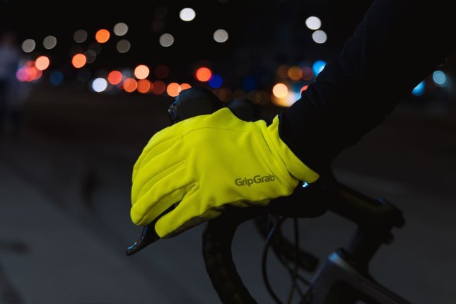 Detailfoto einer Hand in einem neongelben Handschuh der Marke GripGrab an einem Fahrradlenker. Im Hintergrund der nächtlichen Szene sind unscharf bunte Lichter erkennbar.