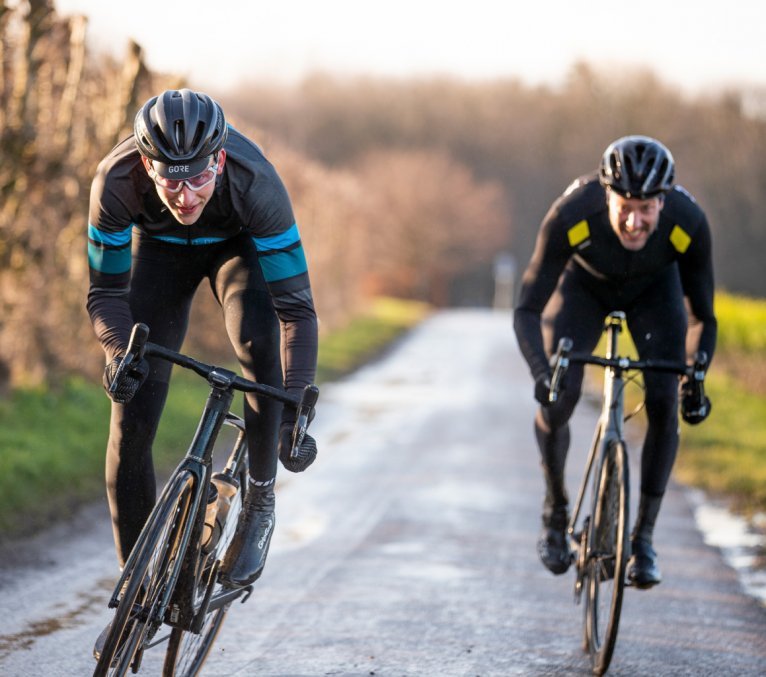 Two cyclists in black sportswear with helmets are riding on a winding road through a forest with autumn-colored trees. The bikes have deep rims and brown tires.