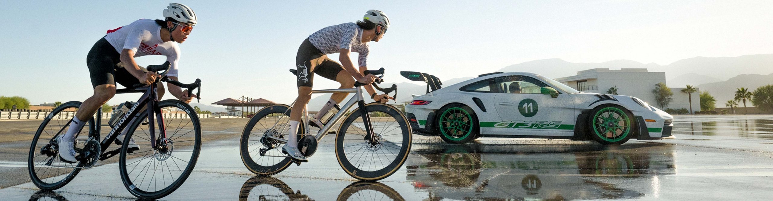 Dos ciclistas de carretera montando bicicletas Cannondale CAAD14 en una pista mojada junto a un coche deportivo Porsche 911 GT3 RS.