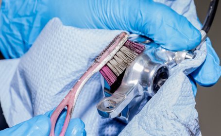 Gros plan d’une personne portant des gants bleus nettoyant un étrier de frein de vélo avec une brosse et un chiffon.