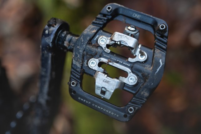 Detail photo: A black OneUp Clip Pedal with a stainless steel cage. In the blur, an autumn forest floor is visible.