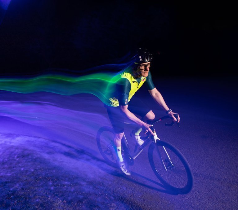 Ein Rennradfahrer mit grellgelber Weste fährt bei Nacht durch einen lilafarbenen Lichtkegel. Er zieht aufgrund der langen Verschlusszeit eine Spur bunter Lichter hinter sich her. 