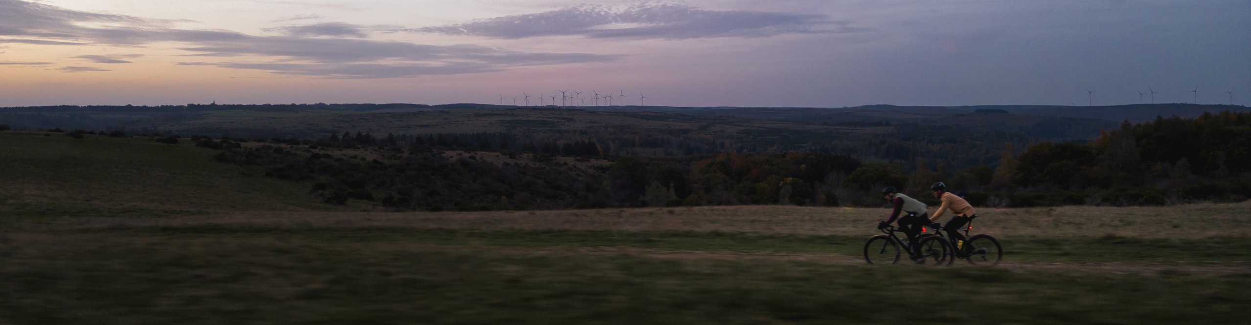 Zwei Gravelbiker fahren in der Dämmerung durch eine offene Landschaft mit Windrädern am Horizont – Fokus auf Ausdauer und Abenteuer im Gelände