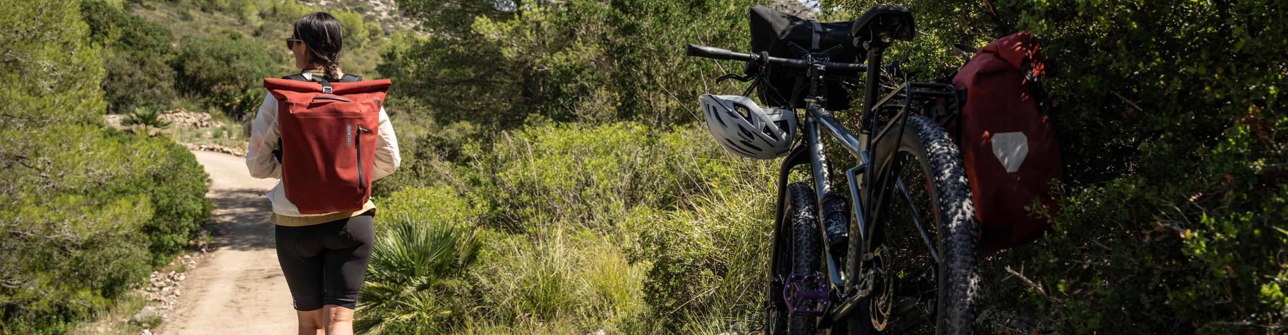 Woman with a red ORTLIEB backpack walking on a nature trail next to a bicycle with an ORTLIEB pannier bag