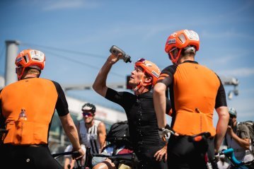 Ciclistas de carretera de bike-components en Rad am Ring hacen una pausa y se refrescan con una botella, con equipación naranja y negra, casco y dorsales.
