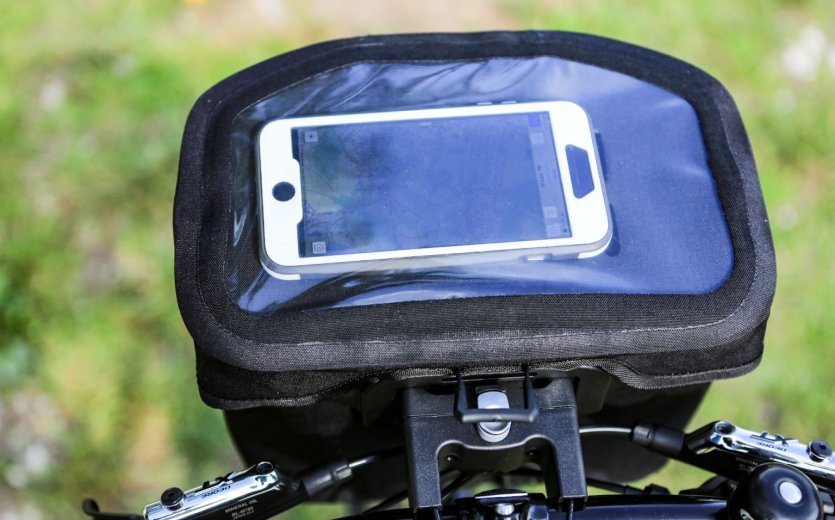 A black bicycle bag with a transparent compartment holding a smartphone is attached to the handlebar of a bike. A blurry green field is visible in the background.