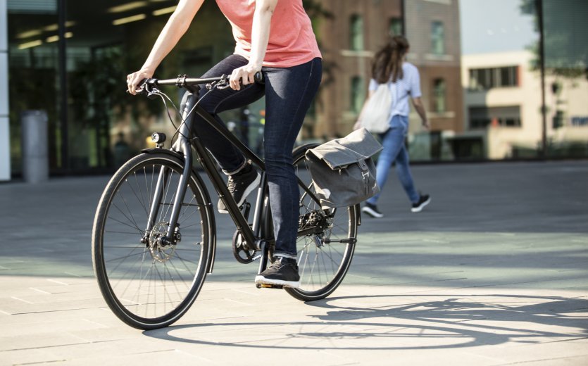 Una persona con una camiseta rosa monta una bicicleta negra con una bolsa de bicicleta gris de Ortlieb sujeta. Al fondo, se ve a otra persona y un entorno urbano con edificios.