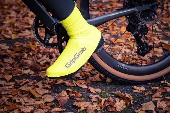Un pie izquierdo dentro de un cubrecalzado ajustado, amarillo neón e impermeable, apoyado en un pedal de bicicleta. El suelo está cubierto de hojas otoñales rojizas.
