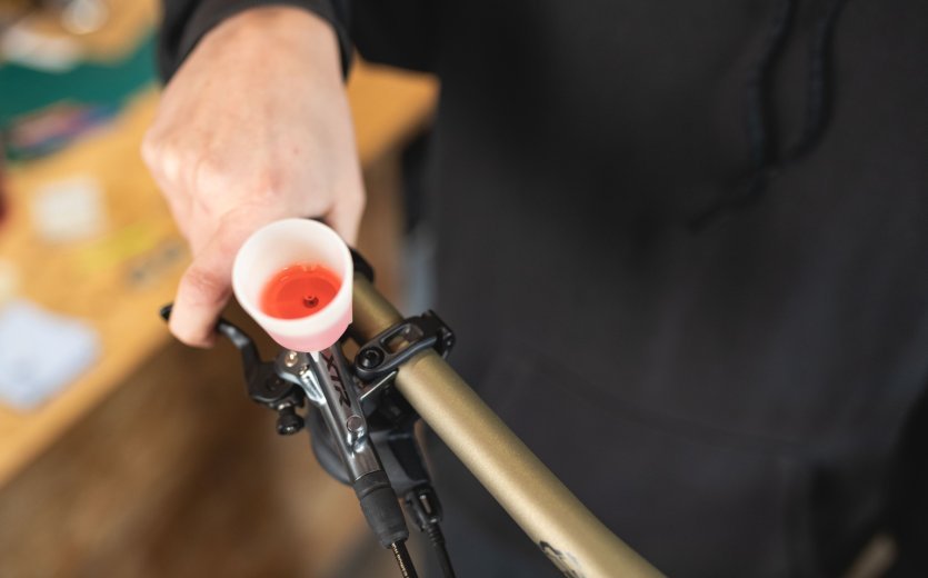 Eine Hand hält einen Bremshebel eines Fahrrads, an dem ein Entlüftungstrichter mit rotem Mineralöl befestigt ist, während der Hintergrund eine Werkstatt mit Holztisch zeigt.
