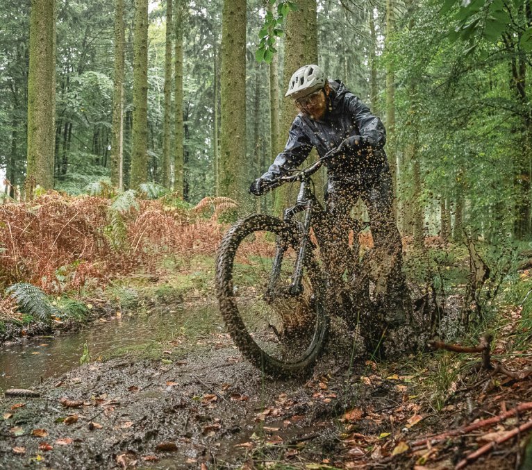 Ein Mann auf einem MTB fährt durch eine große Pfütze im Wald. Matschwasser spritzt in alle Richtungen.
