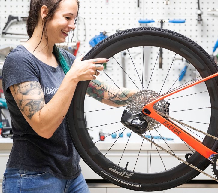 Isa, du service clientèle de bc, monte la roue arrière Zipp 303 sur un vélo de gravel Factor LS orange. Le vélo est suspendu dans un pied d'atelier.