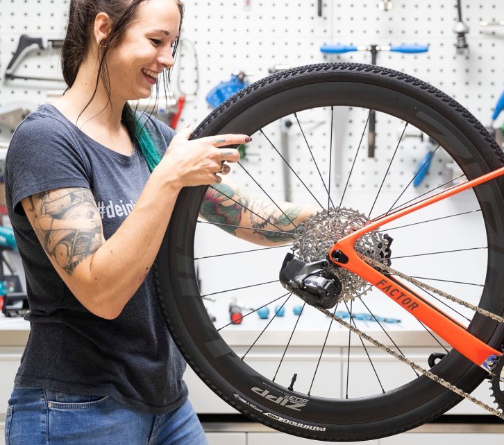 Isa from the bc Service Department mounts the rear Zipp 303 wheel on an orange Factor LS gravel bike. The bicycle hangs on a repair stand.