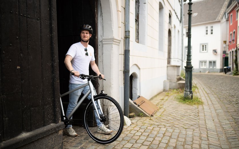 A person stands with a bicycle on a cobblestone street in front of a building in a city, surrounded by historic architecture.