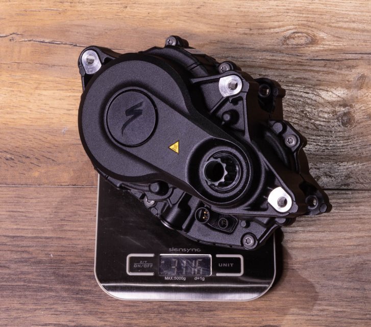 A Specialized 3.1 e-bike drive on a kitchen scale on a dark wooden floor. Display shows 3,116 grams