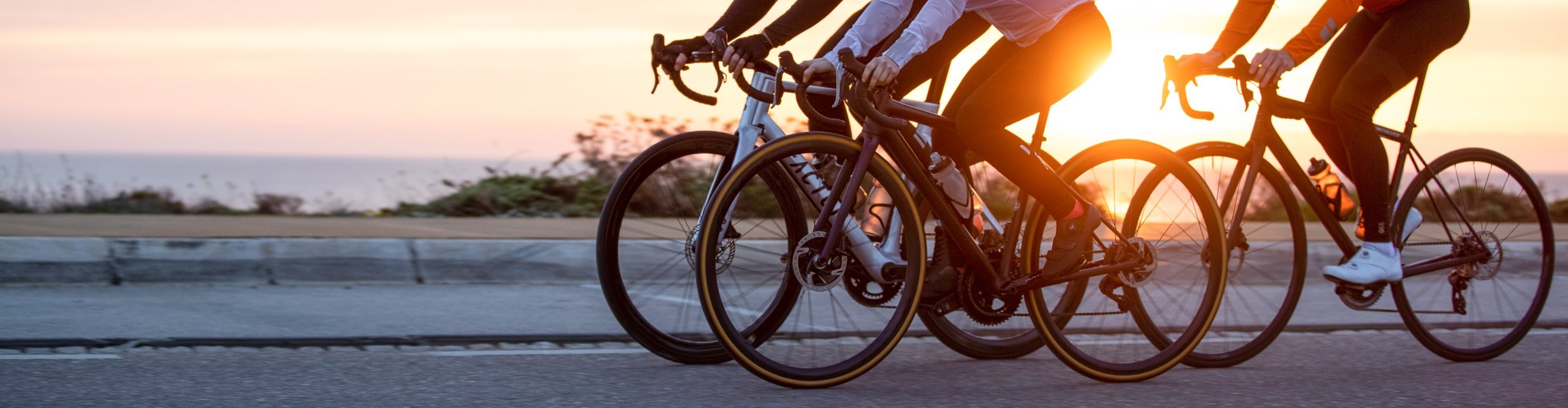 Drei Rennradfahrer fahren bei Sonnenuntergang nebeneinander auf einer Straße und tragen Helme sowie Sportbekleidung – dynamisches Motiv für attraktive Fahrrad Angebote aus allen Kategorien.