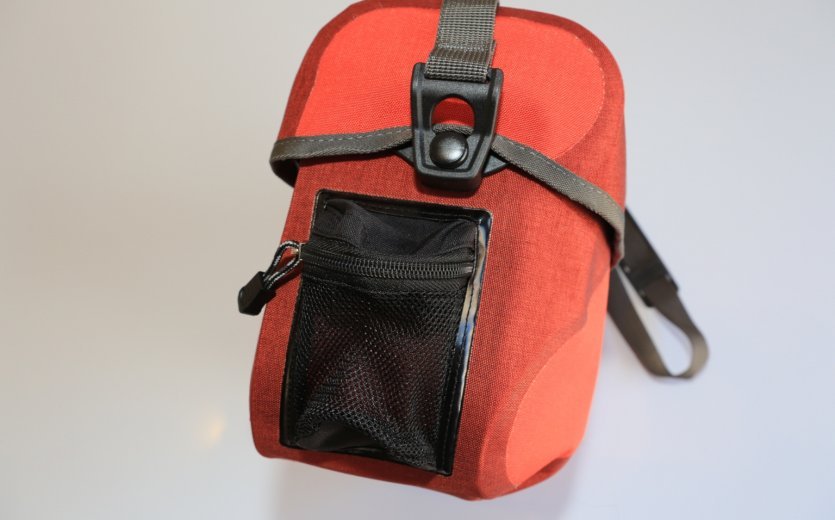 A red Ortlieb bicycle bag with a black mesh pocket on the side and a mount on top.