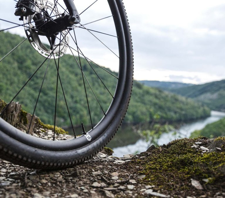 Gros plan de la roue avant d’un vélo gravel sur terrain rocheux avec vue sur une vallée et des collines boisées.