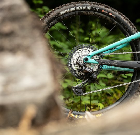 Detrás de una piedra difuminada se ve el lado de la transmisión de una bici de montaña de color turquesa.