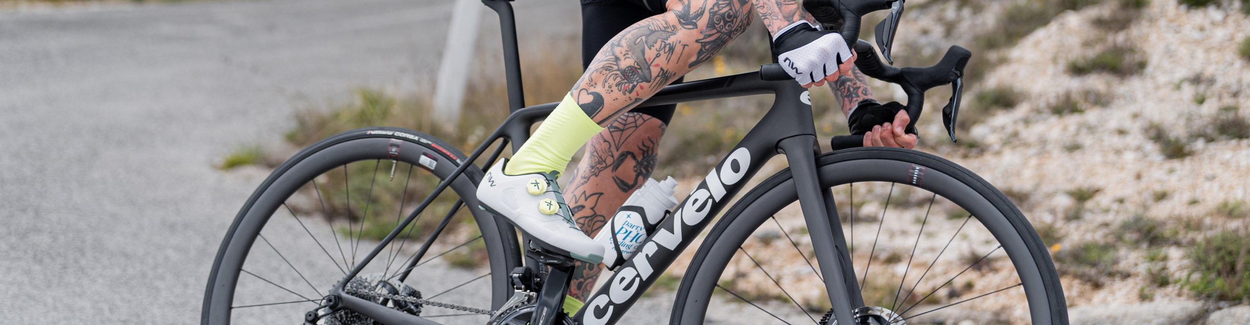 Mann mit Tattoos und Fahrradbekleidung fährt auf einem Cervélo-Rennrad eine kurvige Bergstraße hinauf. Er trägt einen Helm, Sonnenbrille, Radtrikot und neonfarbene Socken. Im Hintergrund sind bewaldete Hügel und ein bewölkter Himmel zu sehen