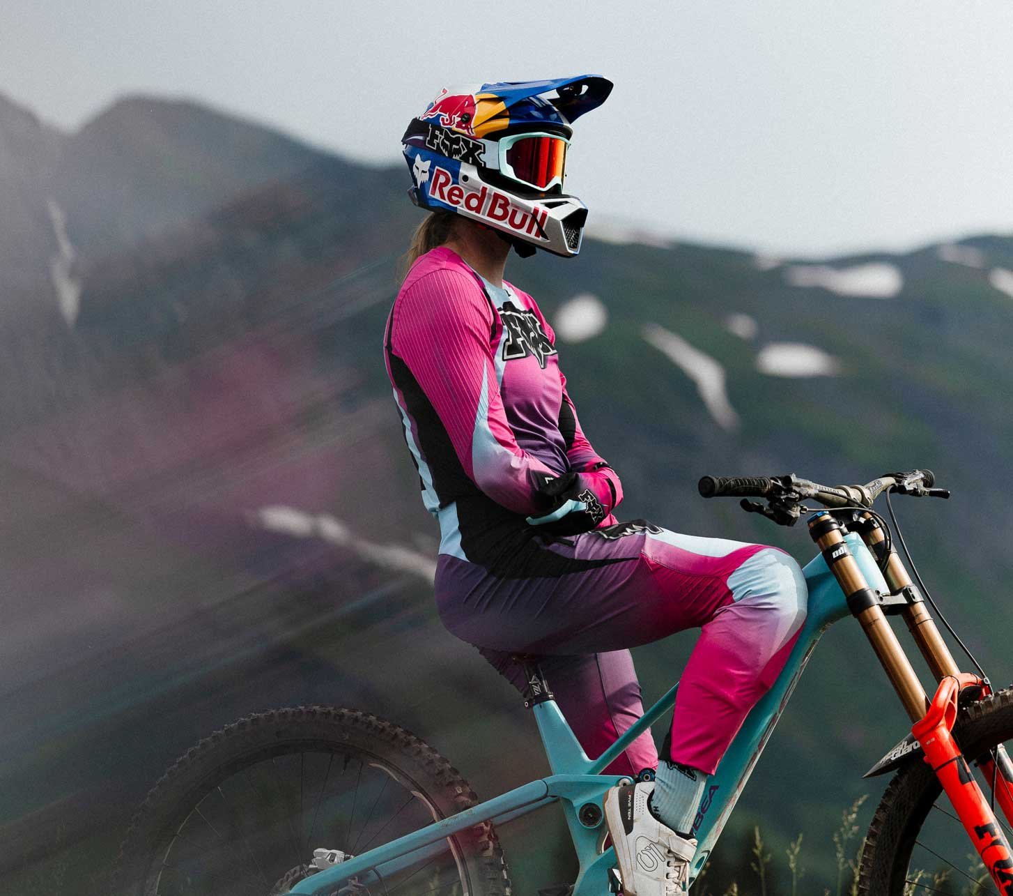 Mountainbikerin mit Fox Racing Protektoren und Fullface Helm sitzt mit ihrem Downhill Bike vor einer Bergkulisse.