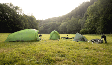 3 VAUDE tents pitched on a meadow surrounded by trees. Next to each tent is a bicycle.