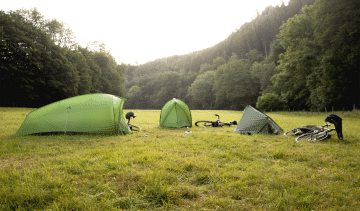 3 tentes VAUDE montées sur une prairie entourée d'arbres. Un vélo est posé à côté de chaque tente.