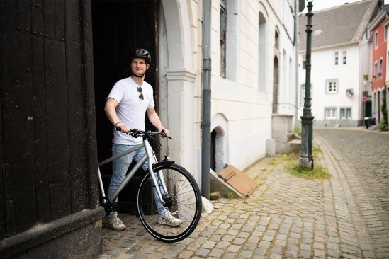 Eine Person steht mit einem Fahrrad in einer gepflasterten Straße vor einem Gebäude in einer Stadt, umgeben von historischer Architektur.