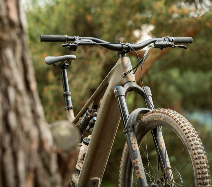 Die Front eines Turbo Levo 4 im Wald. Das Bike wird halb durch einen Baumstamm im Bildvordergrund verdeckt. 