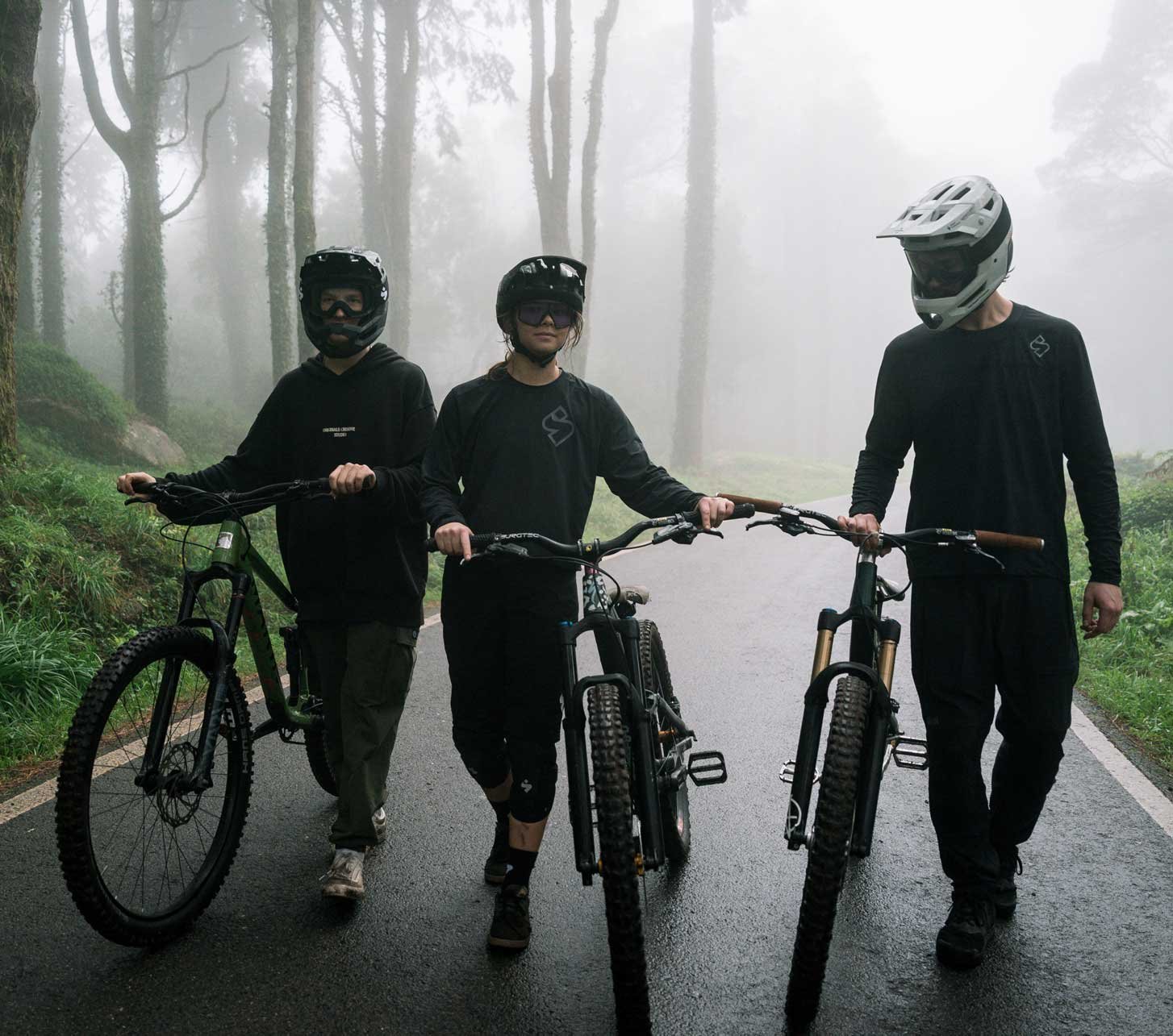 Tres ciclistas de montaña con cascos Sweet Protection empujan sus bicicletas por una carretera mojada en un bosque con niebla.