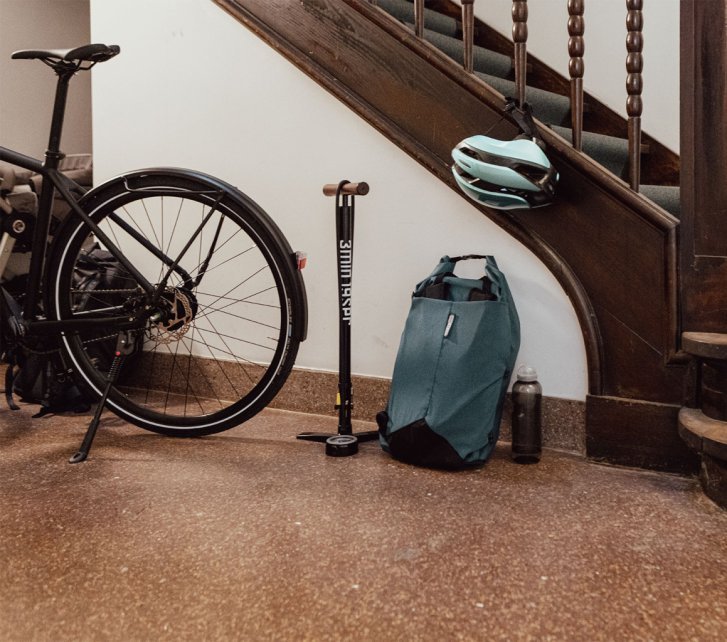 Vélo garé dans un couloir près d’un escalier ; une pompe à pied, un sac à dos, un casque et une gourde sont posés à côté.