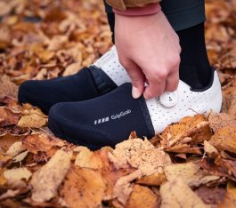 Zwei Füße in weißen Radschuhen stecken in schwarzen Toe-Caps. Im Hintergrund Herbstlaub