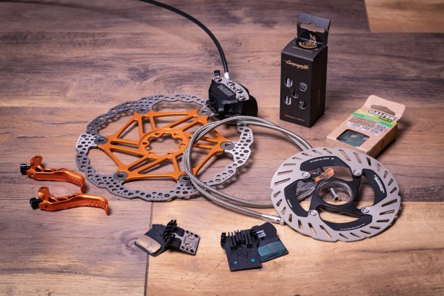 Algunas piezas de mejora para frenos de disco sobre una superficie de madera: discos, latiguillo reforzado, pastillas con aletas y dos palancas anodizadas en naranja.