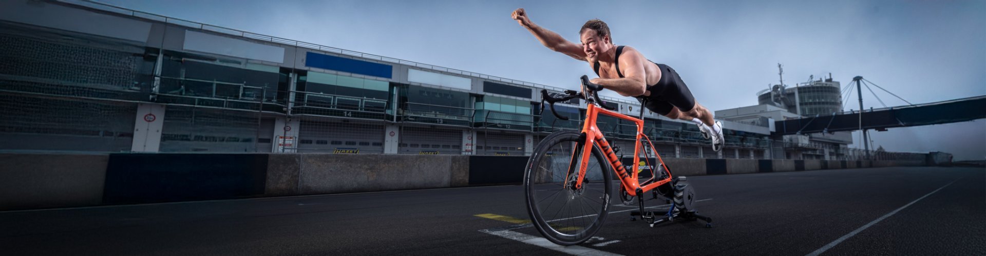 A road bike with a mounted indoor trainer stands on the starting line of a racetrack. A half-naked man lies belly-down on the saddle in a Superman pose.