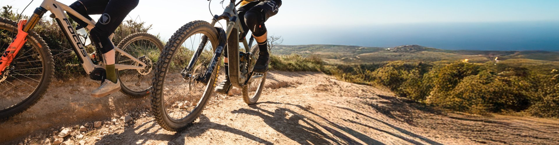 Ein Mann und eine Frau auf E-Mountainbikes fahren auf die Kamera zu. Die Personen sind stark angeschnitten. In. der Entfernung sehen wir einen blauen Ozean.