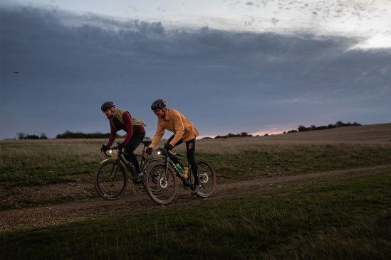 Dos ciclistas con luces en sus bicicletas pedalean por un camino de tierra al atardecer, rodeados de campos y un cielo nublado.