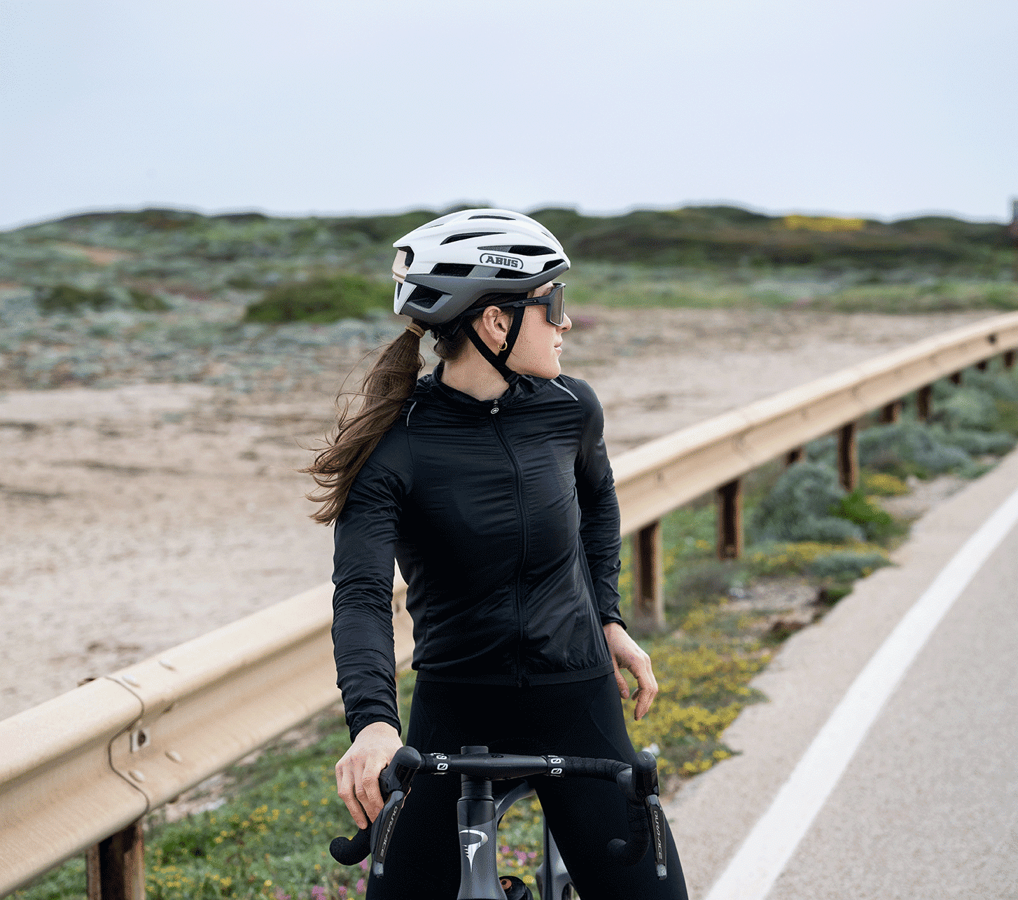 Rennradfahrerin mit ABUS Fahrradhelm steht mit ihrem Rennrad am Rand einer Küstenstraße.