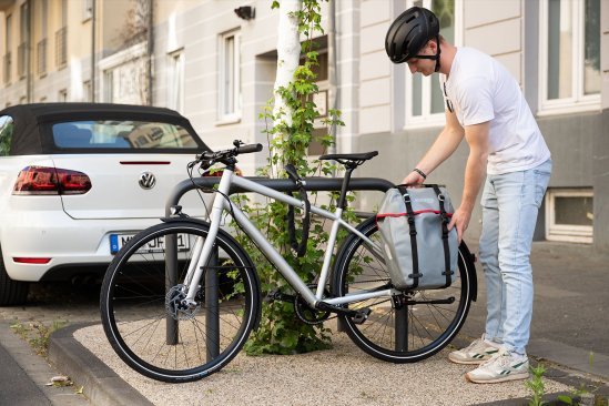 A person locks a bicycle with a pannier bag to a bike rack in an urban setting, next to a parked car and a building with ivy.