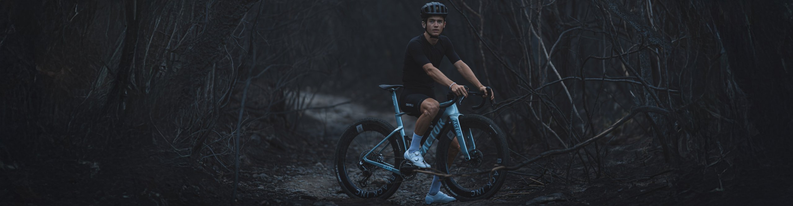 Ciclista de gravel sobre una Factor Elara azul en un camino pedregoso de un bosque oscuro.