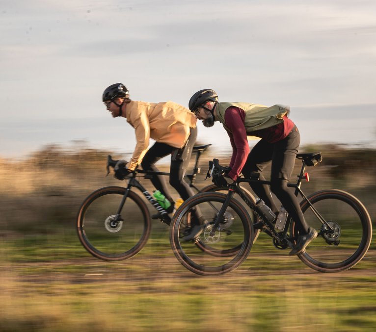 Zwei Gravelbiker rasen nebeneinander über einen Schottertrail, dynamisch in tiefer Rennposition, umgeben von herbstlicher Landschaft – Fokus auf Geschwindigkeit und Gravel-Performance