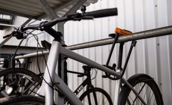 Bicicleta asegurada con un candado en U al cuadro y a una barra metálica en un aparcamiento cubierto.