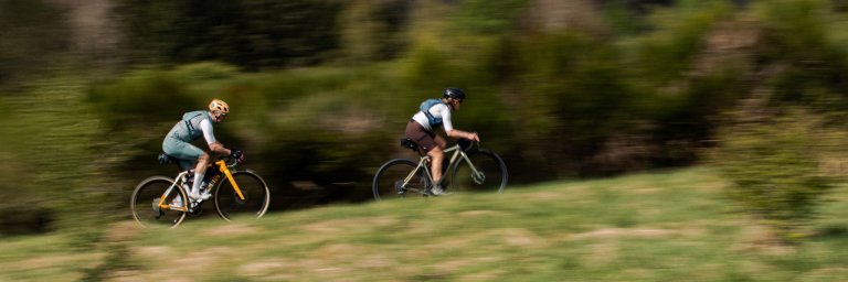 Herbert et Katrin filent sur leurs vélos gravel à travers un paysage naturel verdoyant. Photo filée prise de côté, les cyclistes sont nets, l’arrière-plan est flou en raison du mouvement.