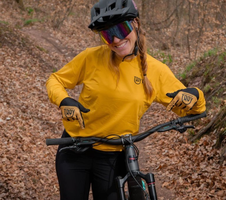 Mujer con maillot amarillo de MTB, casco negro y gafas de colores sonríe y señala su maillot mientras está sentada en una bicicleta de montaña en un bosque otoñal.