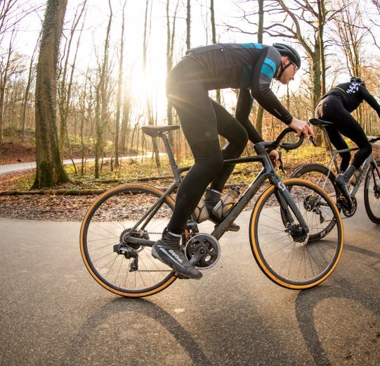 Zwei Rennradfahrer, unterwegs auf Bikes von Scott und Factor, fahren bei herbstlichem Wetter bergauf.