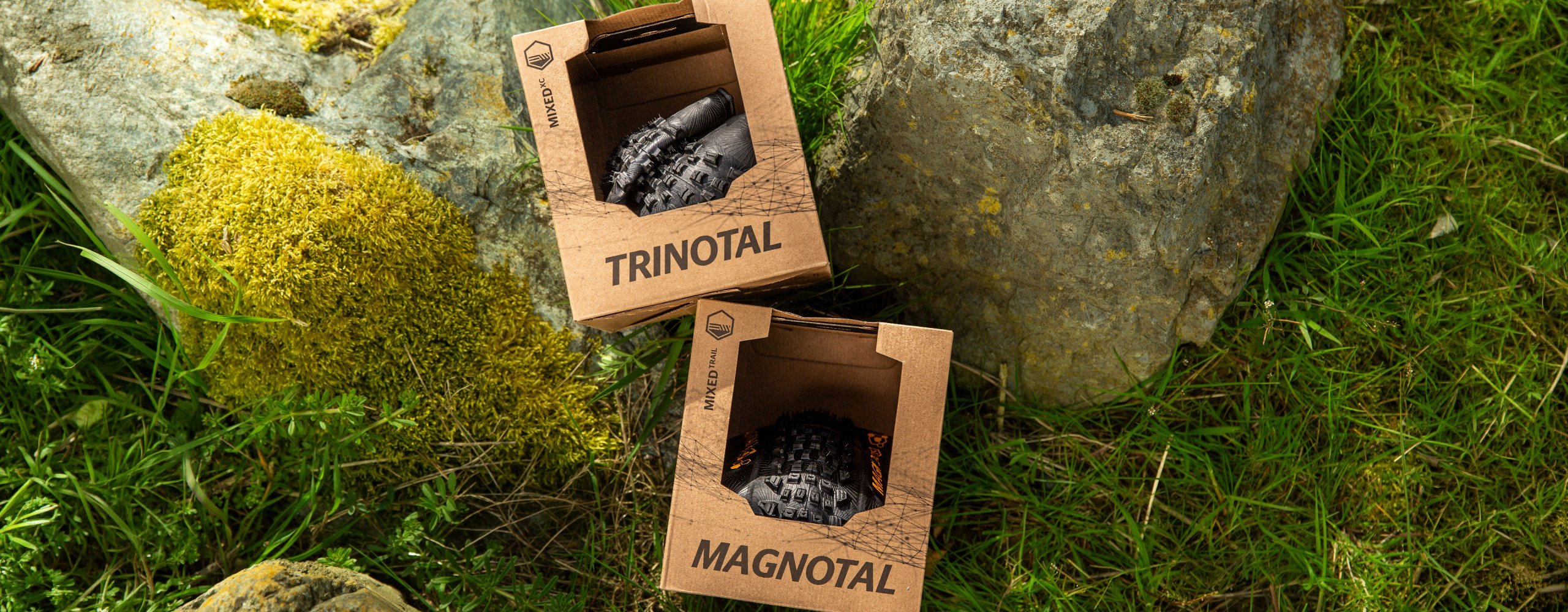 Zwei Continental Fahrradreifen in Kartonverpackungen mit den Namen Trinotal und Magnotal liegen zwischen Steinen und Gras im Outdoor-Setting.