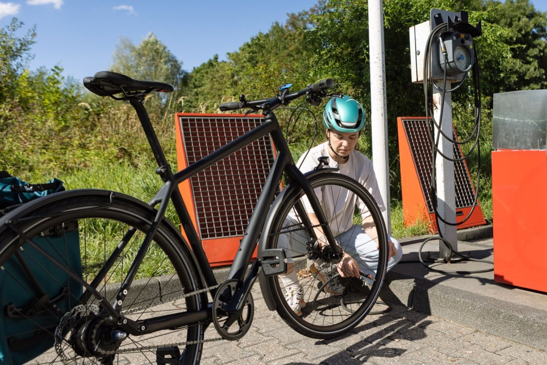 A young woman crouches behind a black city bike and connects the hose of a petrol station compressor to her tyre valve.