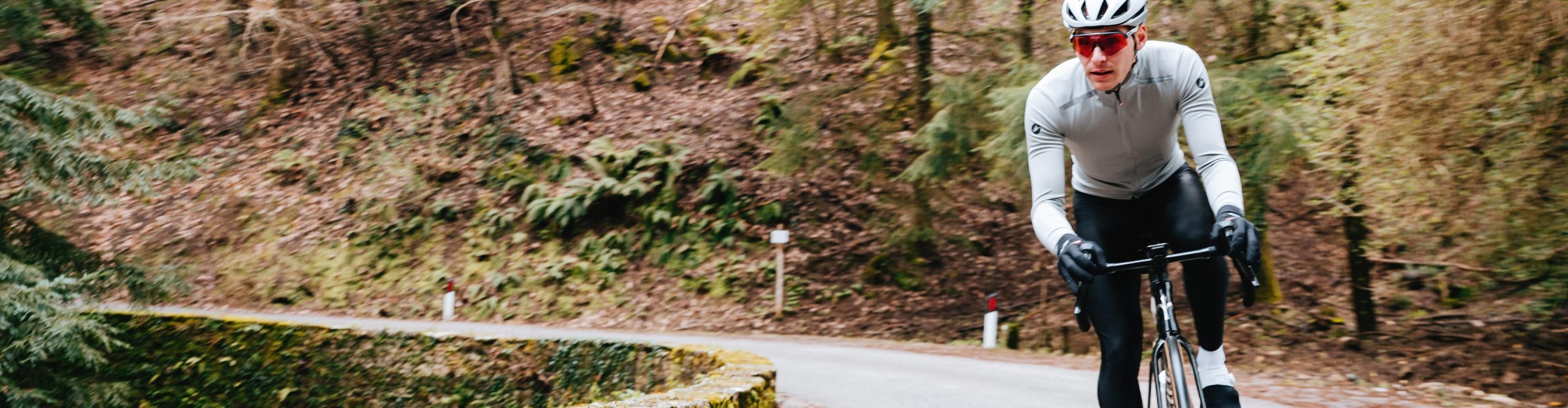 Cycliste sur route en vêtements Castelli pour l’automne et l’hiver roulant sur une route sinueuse en forêt.