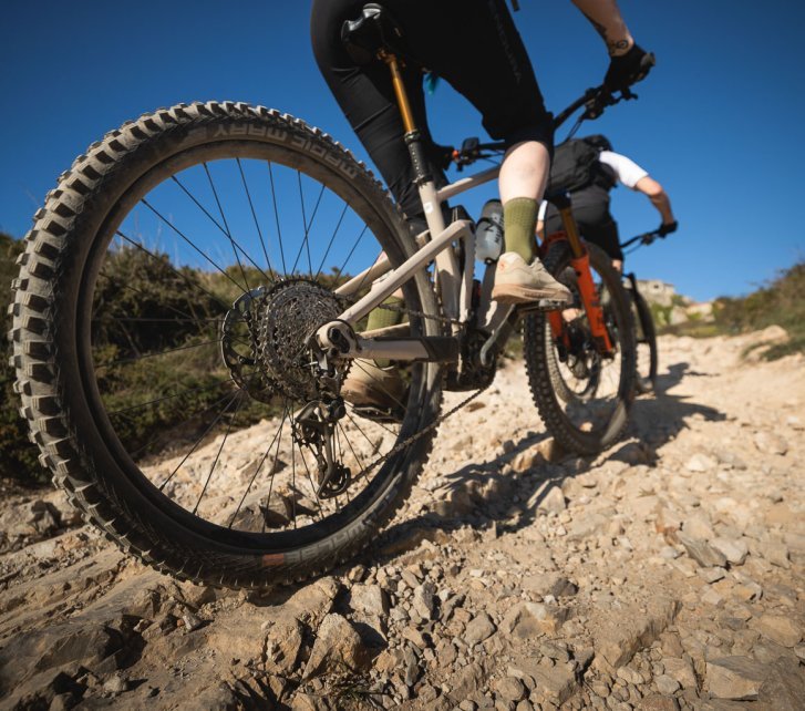 Zwei E-MTB kraxeln einen felsigen Trail hinauf. Die Personen sind star kangeschnitten, Fokus liegt auf dem Hinterrad und Antrieb des hinteren Bikes. Der Himmel ist kräftig blau.