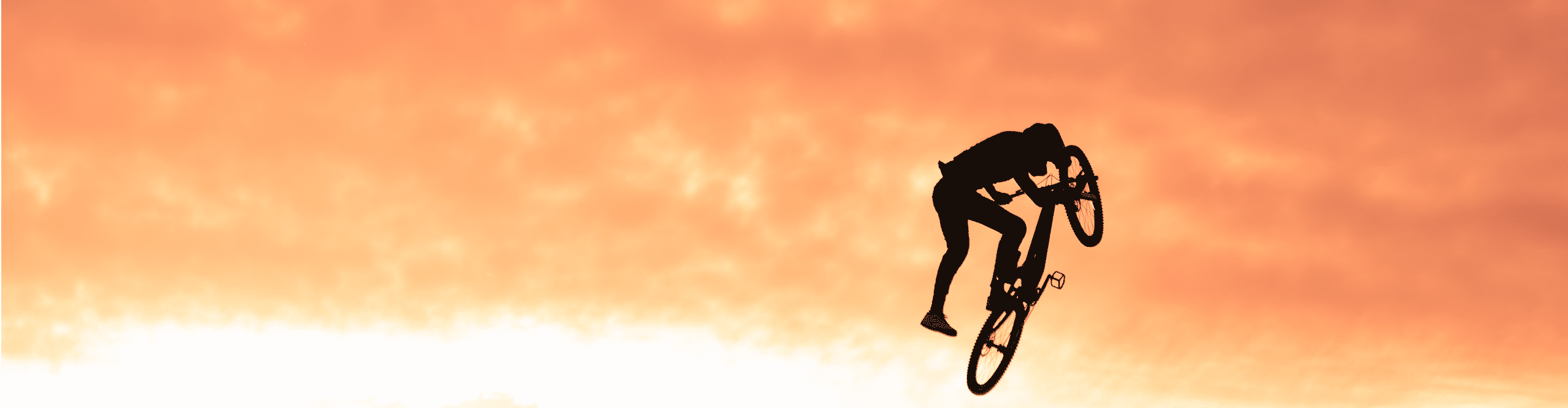 Silhouette d’un vététiste réalisant un grand saut dans un bikepark au coucher du soleil avec des bosses en terre.